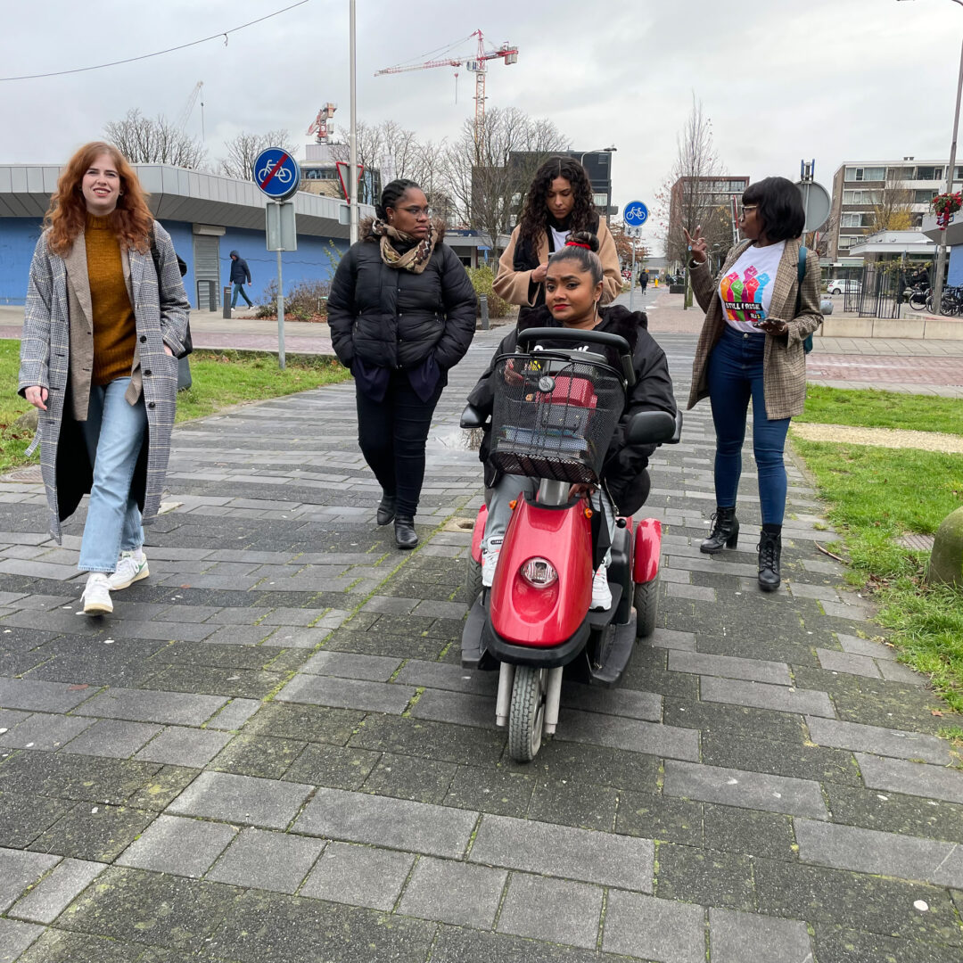 Van links naar rechts; Susan, Manuela, Julia, Jeanette en Anraleida lopen in Voorhof. Julia en Anraleida hebben een BIJ1 t-shirt aan, Jeanette rijdt op haar scooter.